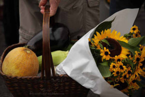 a shopping basket filled with flowers and produce carried by a man at a farmers market