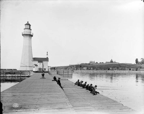 A vintage photo of Oswego's west pier, 1880 stone lighthouse, and Fort Ontario in the background.