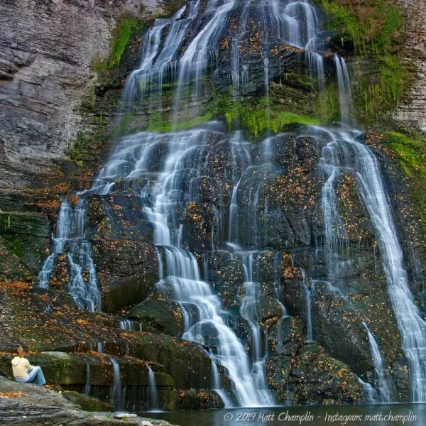 A closeup of Frontenac Falls at Camp Barton in low flow. Fall leaves are sprinkled upon the wet bare rocks of the cliff face. A woman sits in front of the falls.