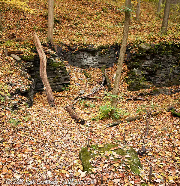 Remnants of an old mill on Catlin Mill Creek at Deckertown Falls. Some stonework is seen along the cliff and metall beems protruding from a large stone in the foreground. The rest of the scene is covered in fall leaves.