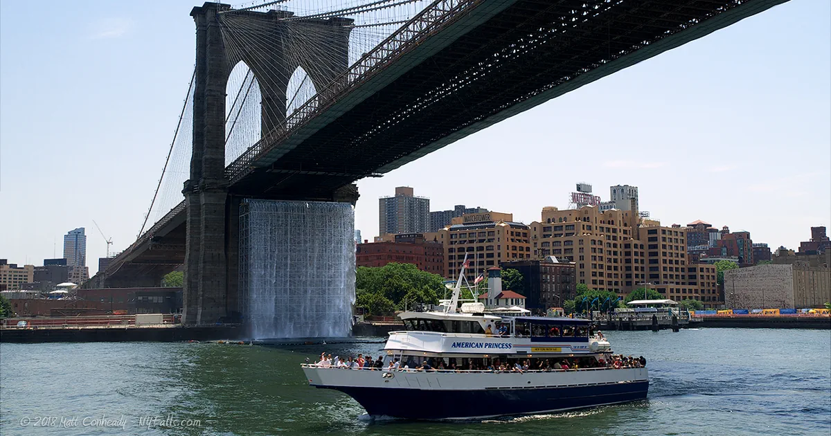 A NYC water taxi passing by a waterfall under the Brooklyn Bridge