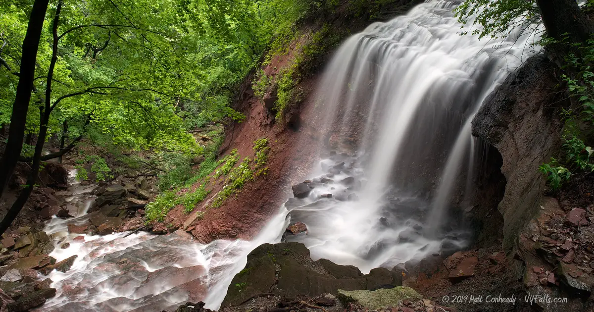 A view of Norton's Falls in high flow. The falls comes out of a tunnel, down a short cascade, and freefalls before turning and tumbling the rest of the way into the Genesee gorge.