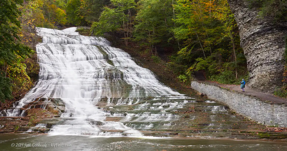 A wide view of Buttermilk Falls, in autumn. someone is standing on the trail leading up the falls.