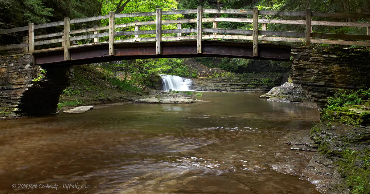 A steel and wooden bridge that crosses Buttermilk Creek. A small waterfall is seen in the distance.