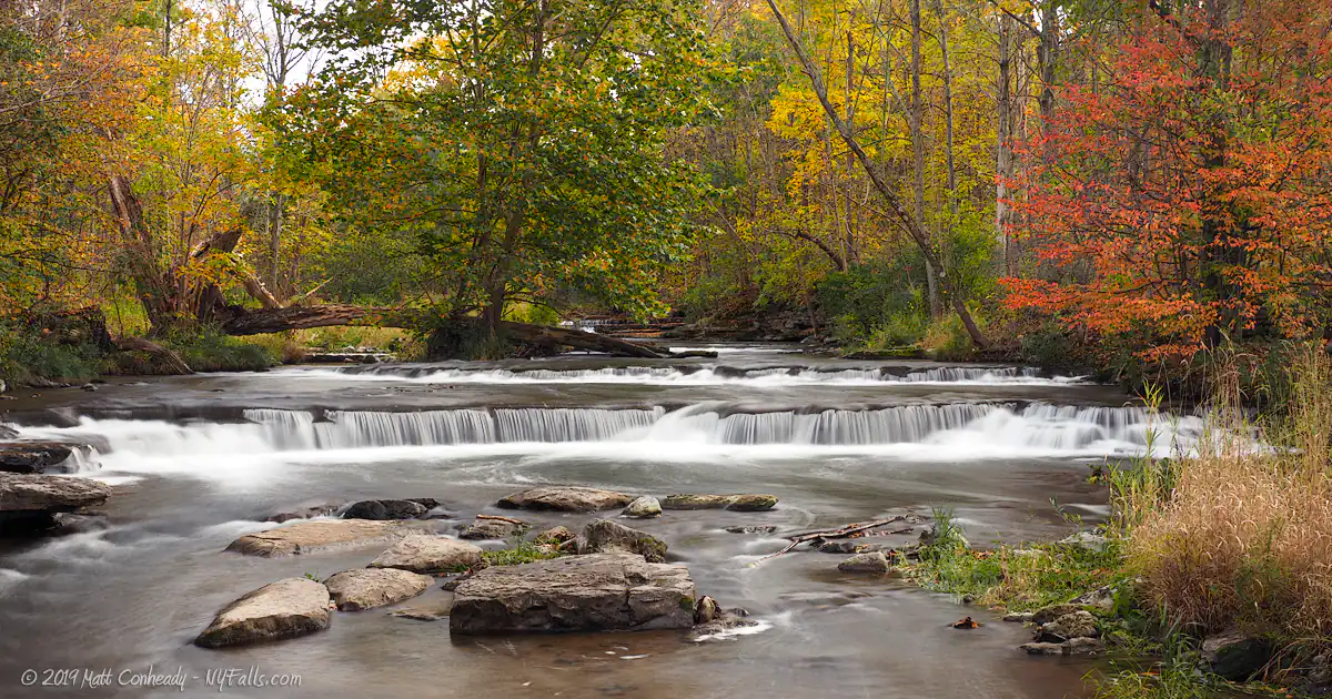 An autumn scene showing a short, but wide waterfall on Chittenango Creek upstream from the falls in a wooded area.