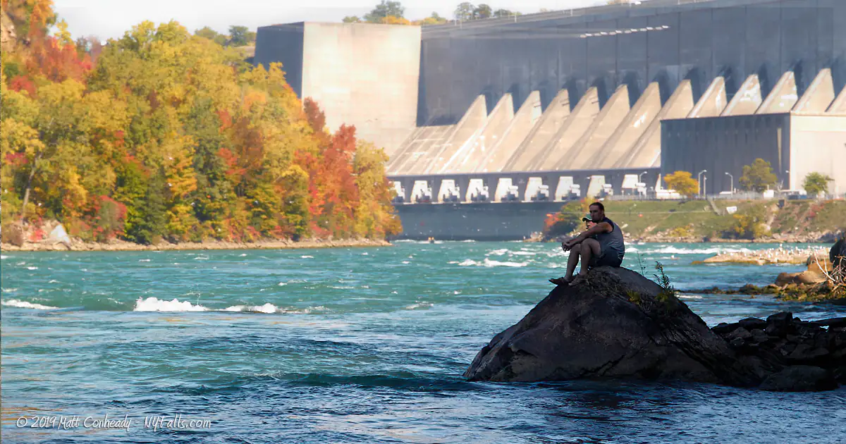 A couple sitting on a large boulder on the Niagara River with the Robert Moses Power plant in the background