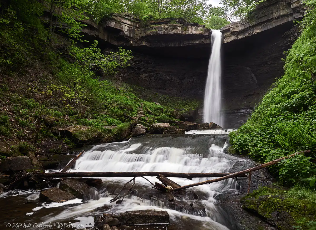 A wide view of upper Carpenter Falls. The freefall leads to s wider, shorter cascade with stones and tree branches stuck in the lower cascade.