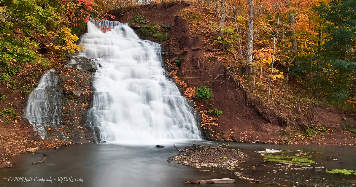 An autumn view of Holley Canal Falls. The surrounding dirt is red, which matches the foliage nicely. A small cascade has branched off of the left of the falls.