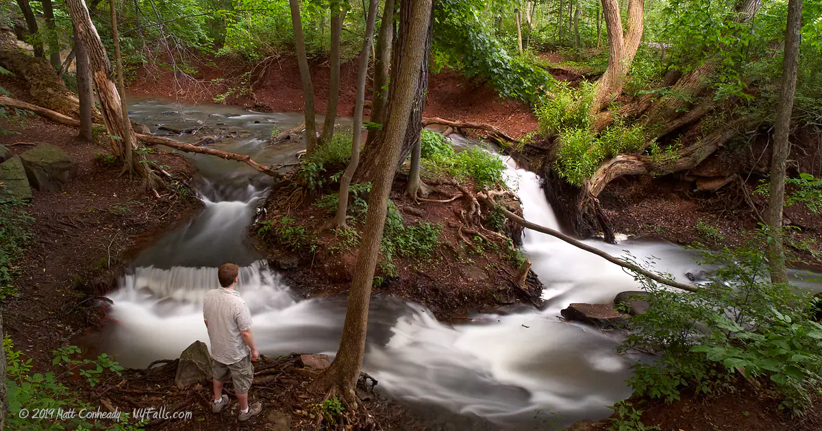 A man stands next to some small waterfalls in the woods above the falls. The dirt is a iron red color.