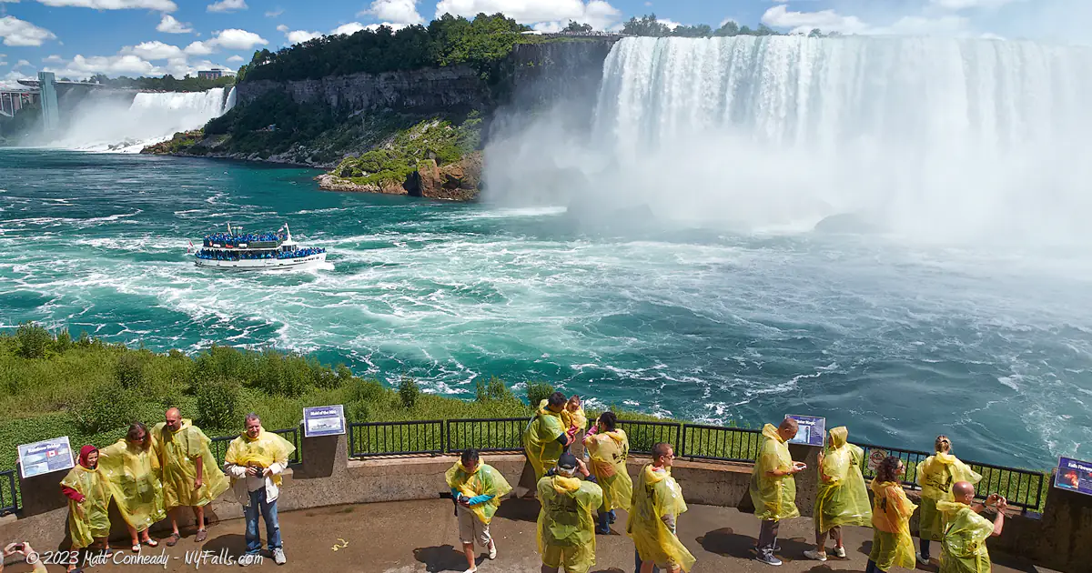 A view of Niagara Falls (American Falls on the left and Canadian Falls in the right) and the Maid of the mist with tourists at the Journey Behind the Falls attraction in the foreground.