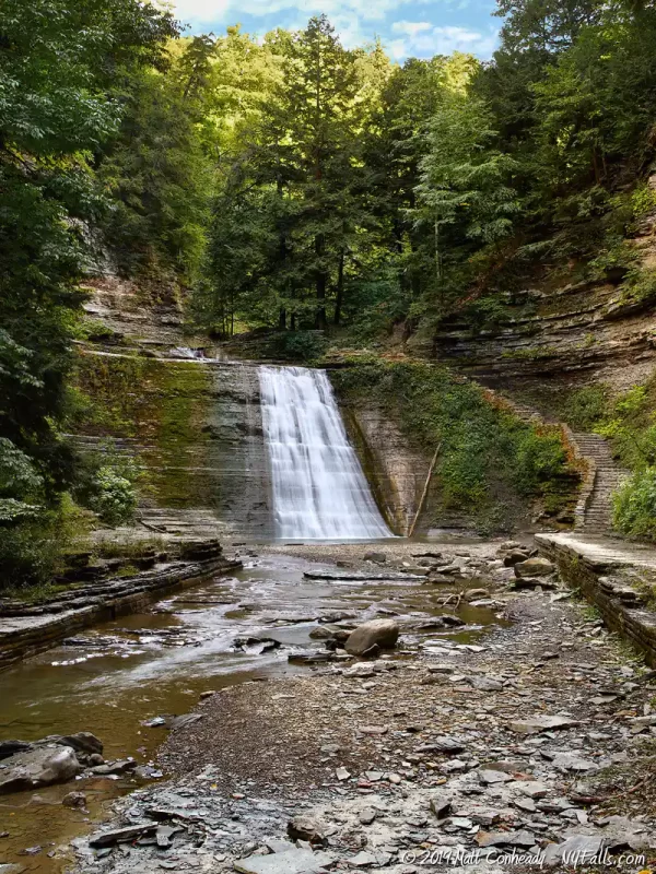 A wide view of a tall waterfalls, with the gorge trail on the right and dense woods just beyond the falls.