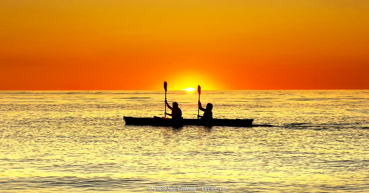 Two people in a kayak paddle past the sunset over Lake Ontario