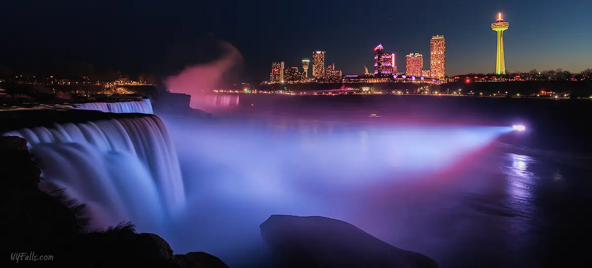 Niagara Falls illuminated in purple with Niagara Falls, Ontario, Canada in the background.