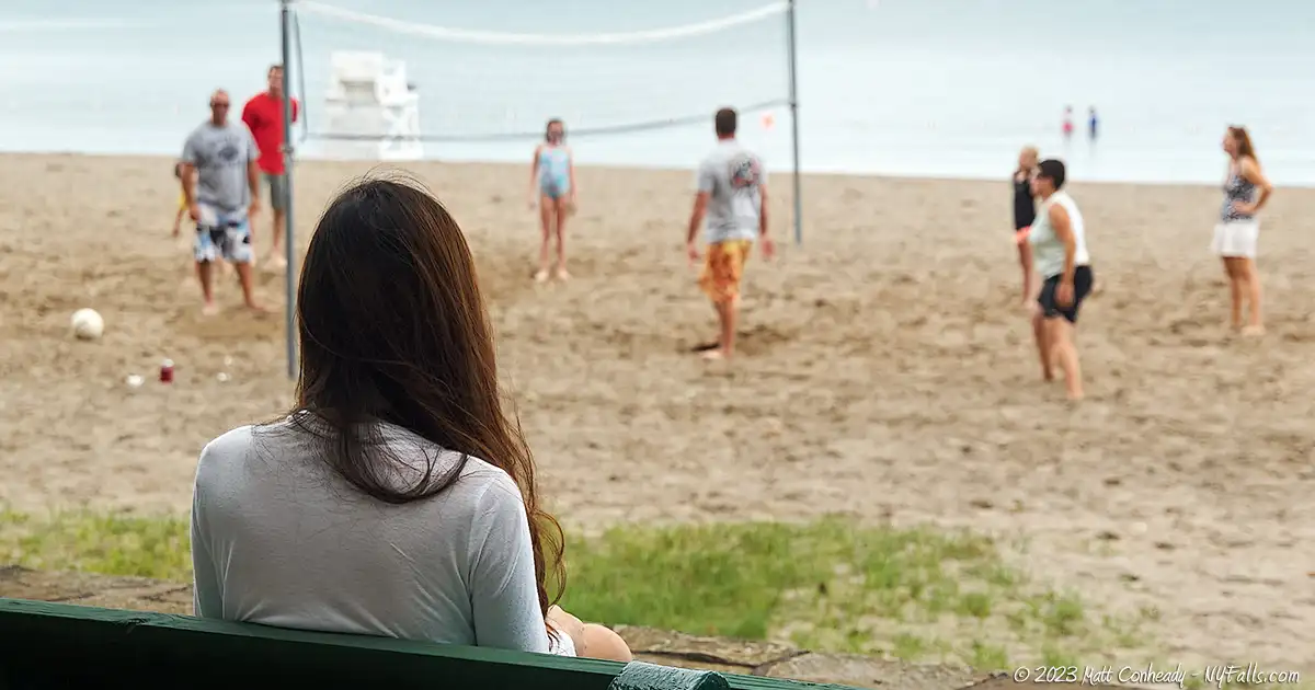 A girl sits on a bench and watches a beach volleyball game.