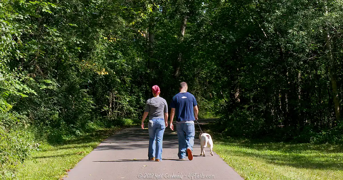 A couple walks a dog on a paved path in Onondaga Lake Park