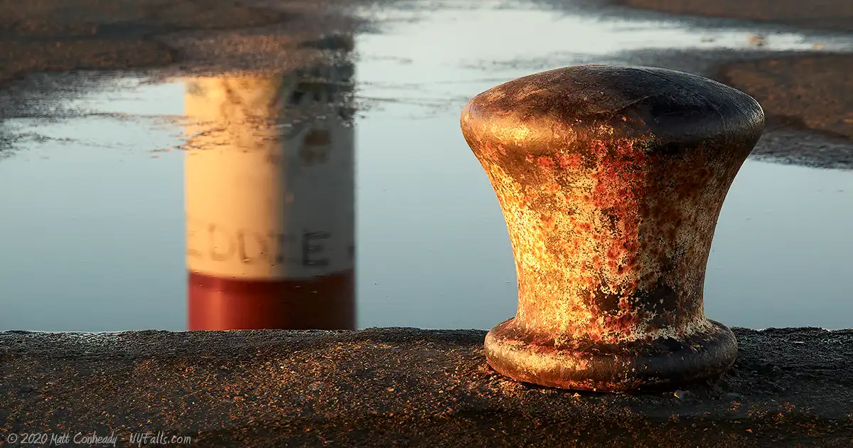 A close up of boat cleat on the Charlotte Pier with a reflection of the beacon, or "lighthouse" in a puddle