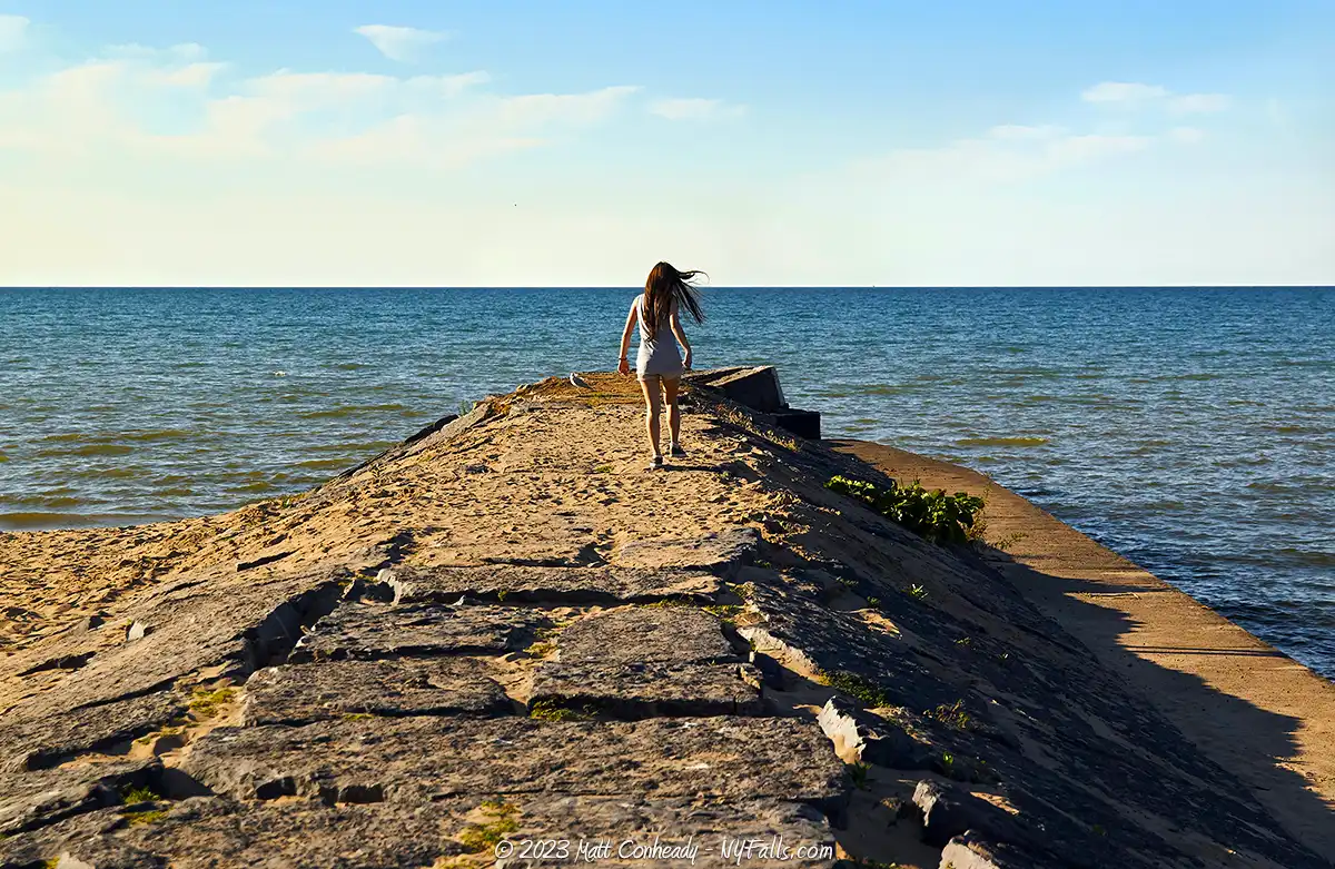 A woman walks down a concrete pier towards Lake Ontario in the late afternoon sun at Fair Haven Beach State Park.
