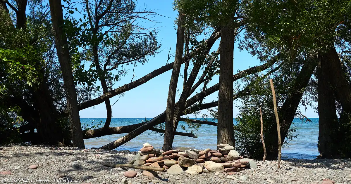 Looking through the trees along the beach at Whistlewood Park. In the foreground someone set up a campfire. In the background is a moderately calm Lake Ontario.