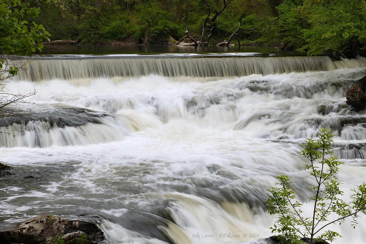 The top portion of the falls which is a broad dam. The natural waterfall is in the foreground.